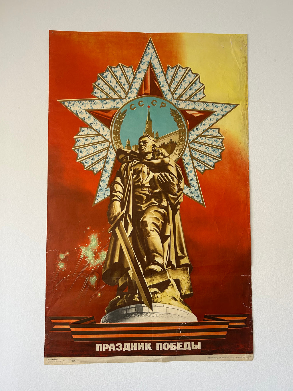 A vintage Soviet Victory poster featuring a gold-toned statue of a warrior holding a sword, set against a large silver and red star emblem with a blue central medallion. The background is a warm red and yellow gradient, with Russian text at the bottom reading 'ЗОННОТНОЕ ЯЗОЙНИЕ'.