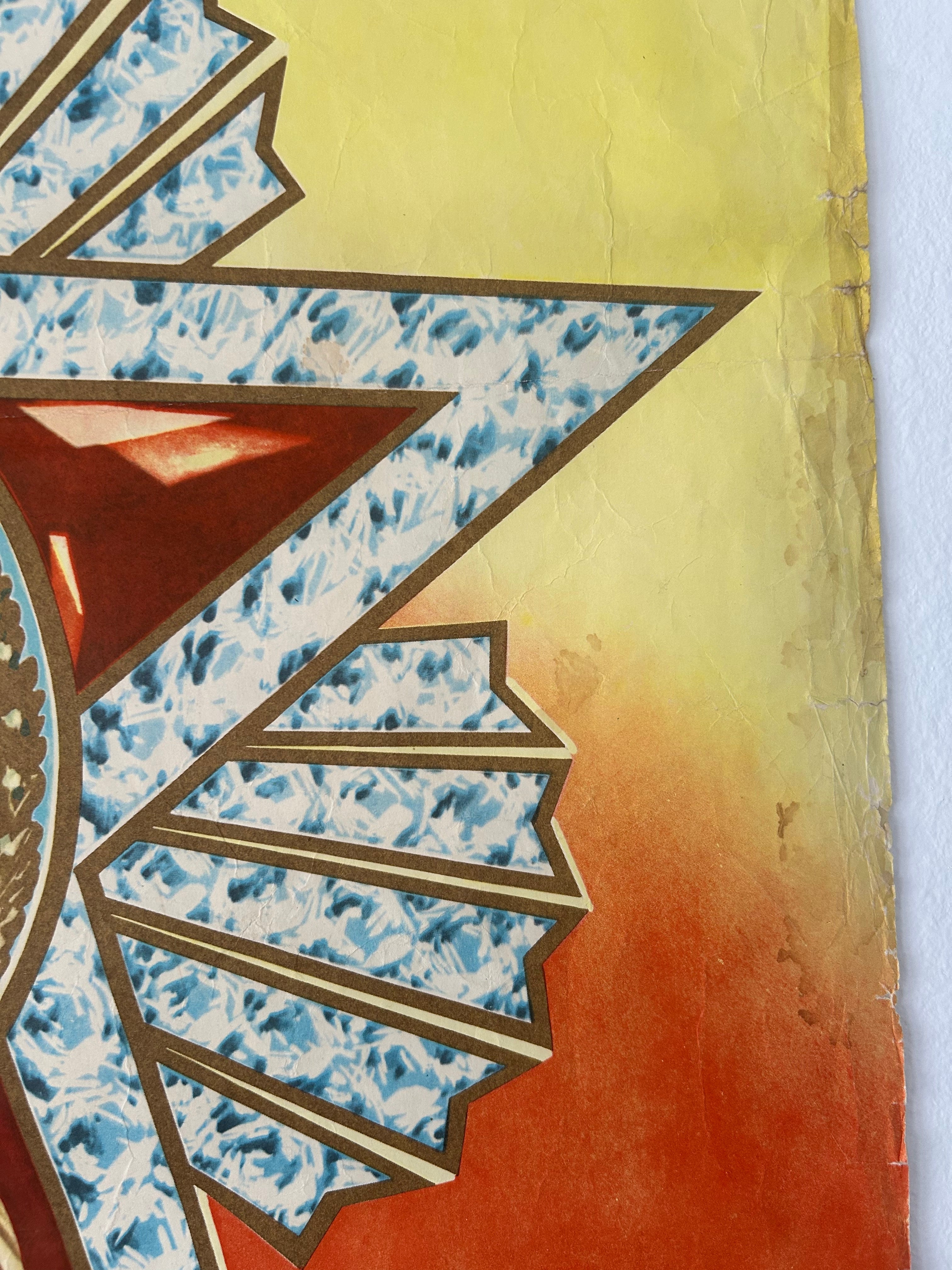 A close-up of a decorative textile or art piece featuring a stylized sunburst motif with blue marbled patterned sections, brown outlines, and a warm yellow-to-orange gradient background.