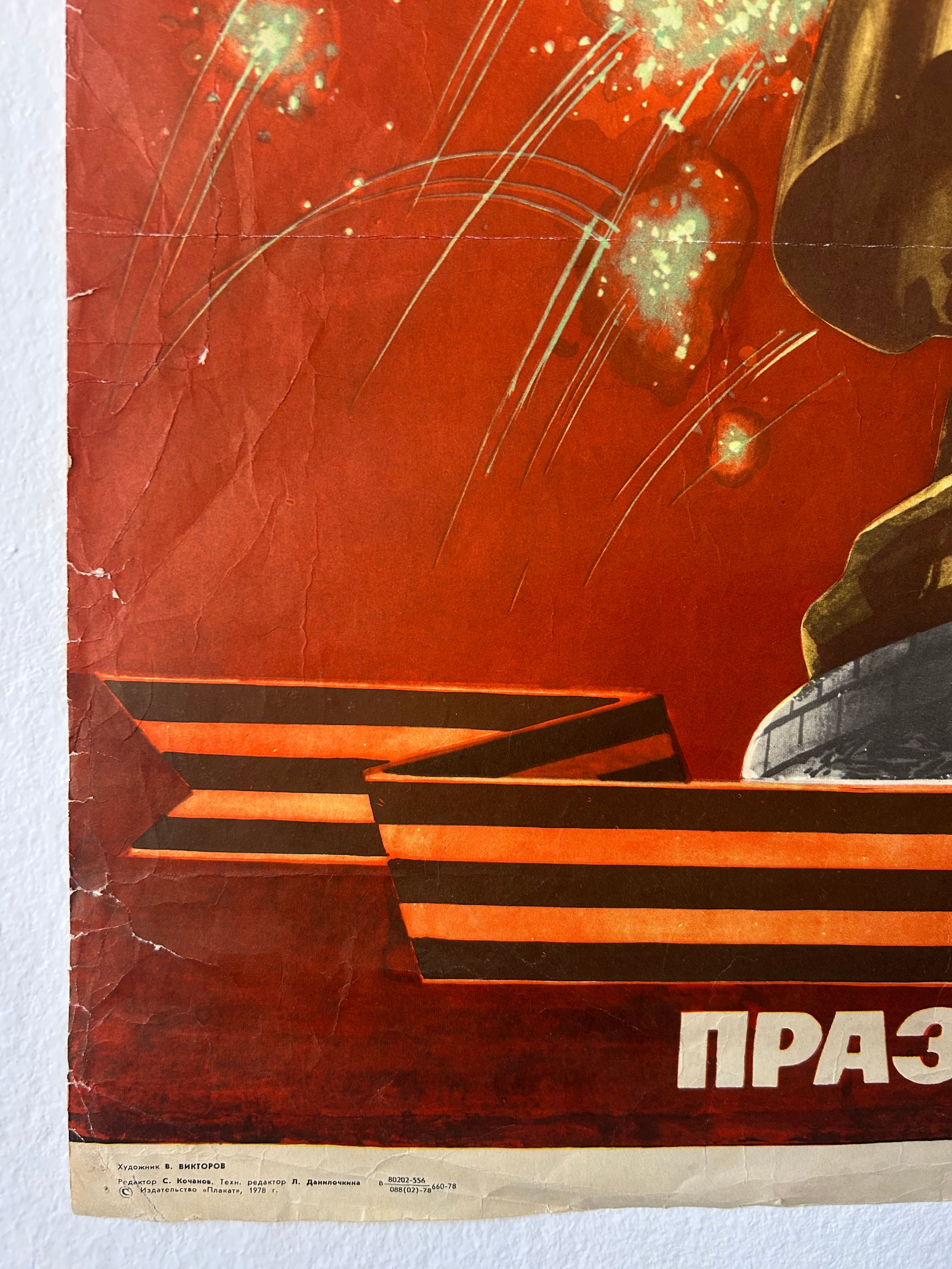 A vintage Soviet propaganda poster print featuring a red background with fireworks, a golden hammer and sickle, and a striped banner with Cyrillic text at the bottom, photographed against a white wall.