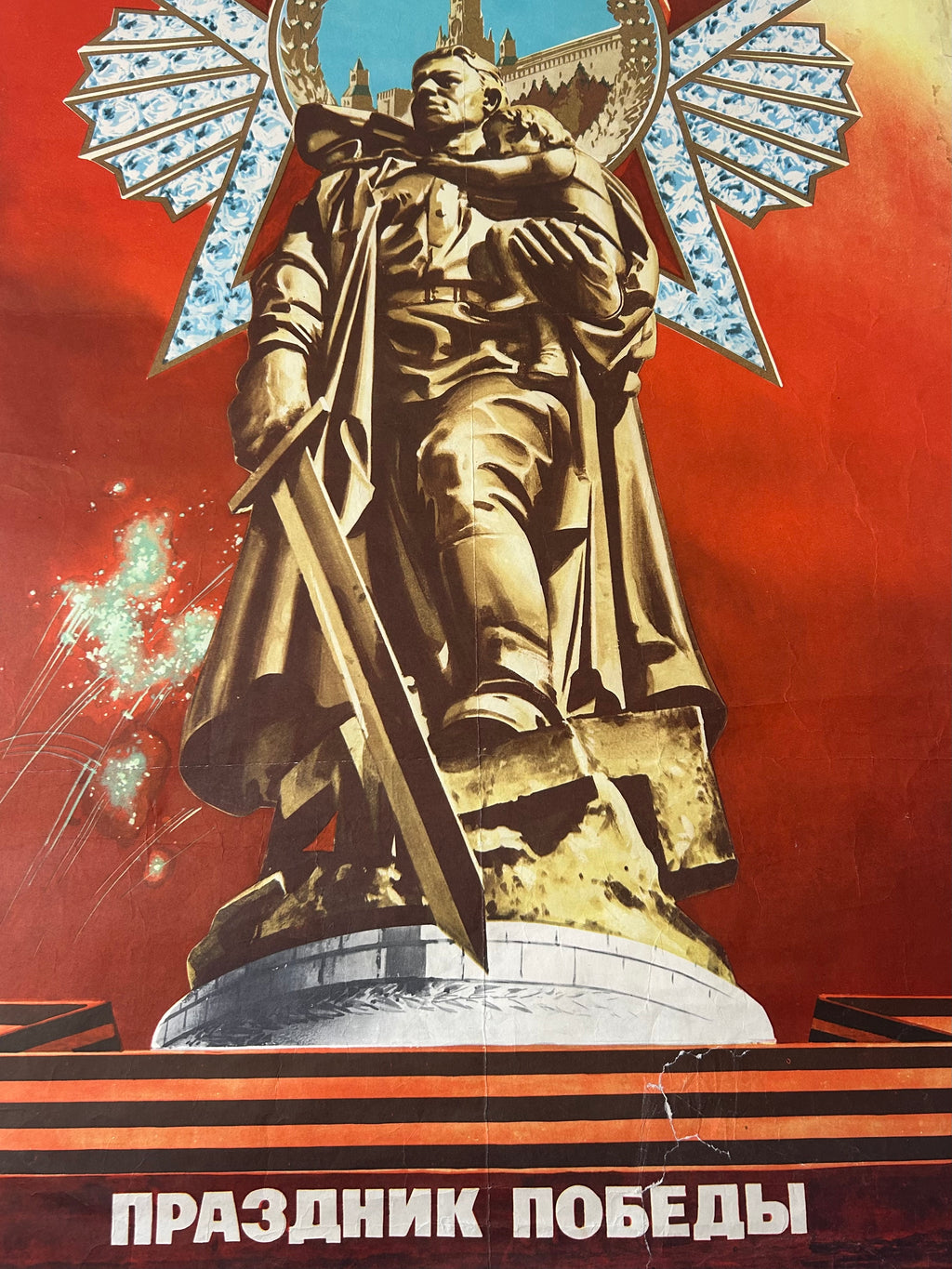 A vintage Soviet-style poster featuring a large golden statue of a soldier holding a sword, set against a vivid red background with a blue sky and white Cyrillic text at the bottom reading 'Prazdnik Pobeedy'.