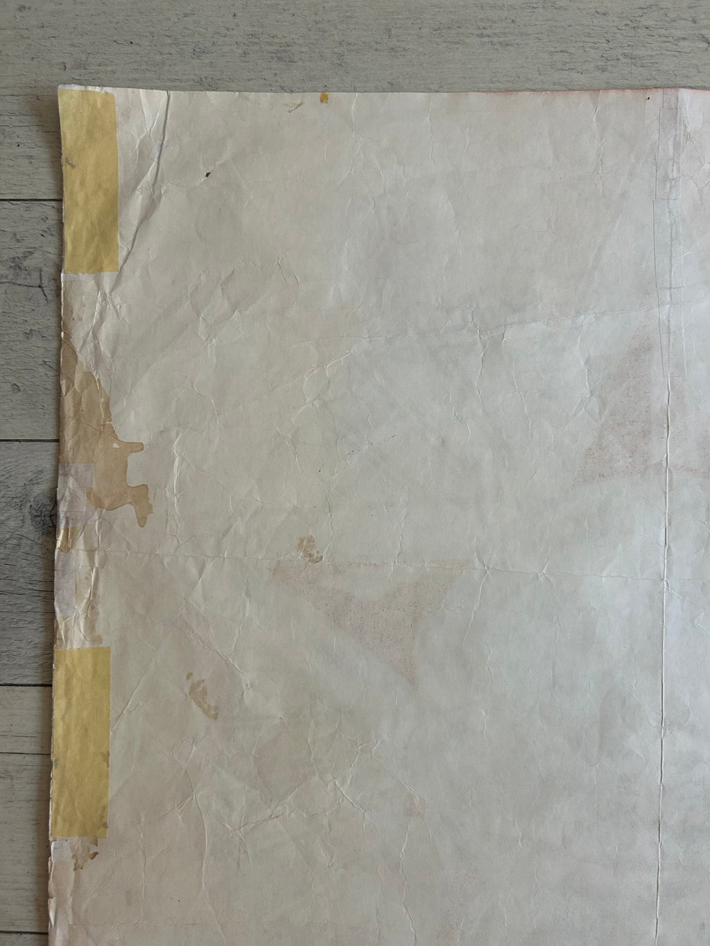 A large, crumpled sheet of off-white paper with yellow tape secured along the left edge, laid flat on a gray wood plank surface.
