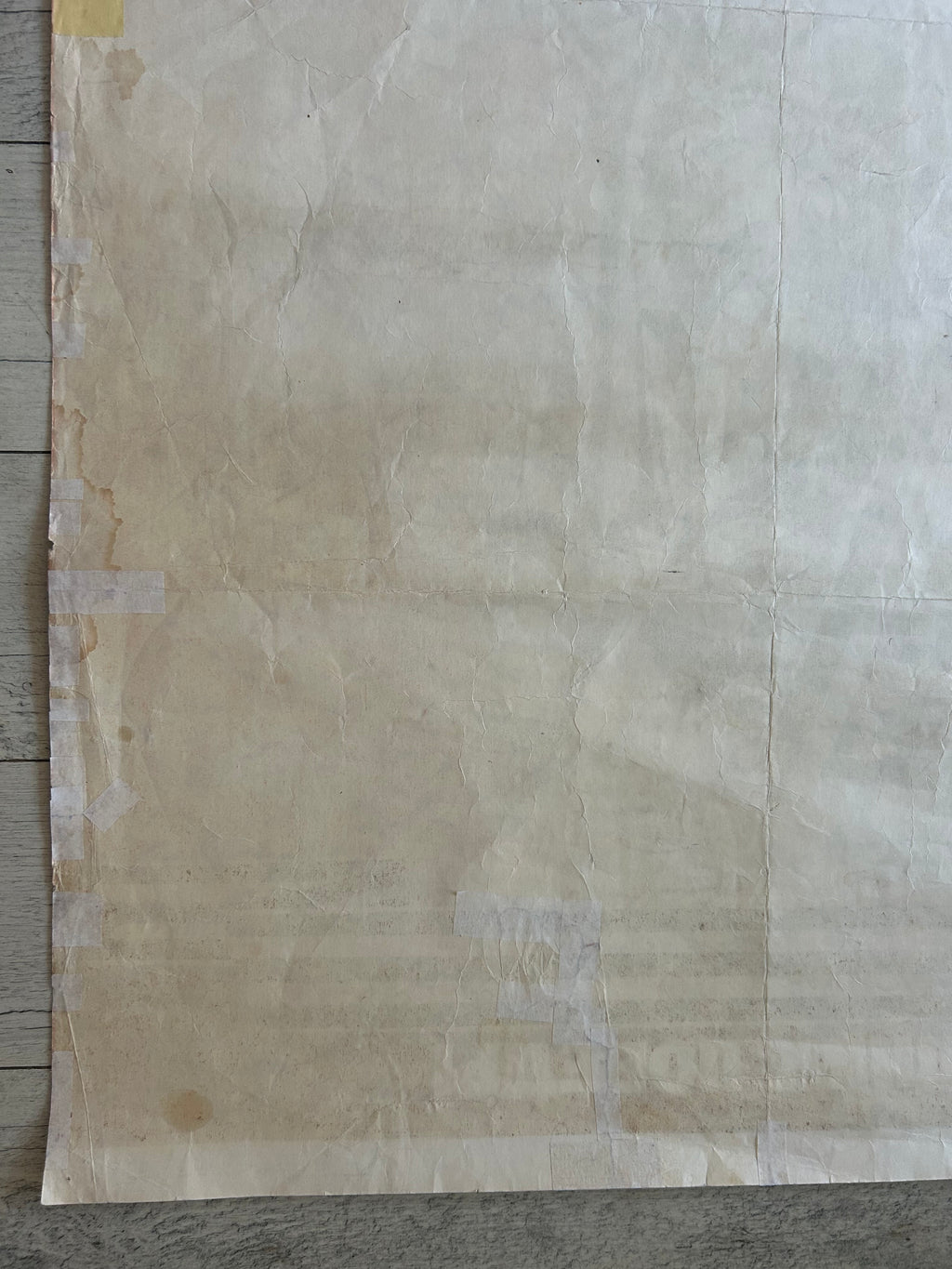 A large sheet of textured, off-white to beige handmade paper with visible creases and a rustic, vintage appearance, taped down on a gray wood floor.
