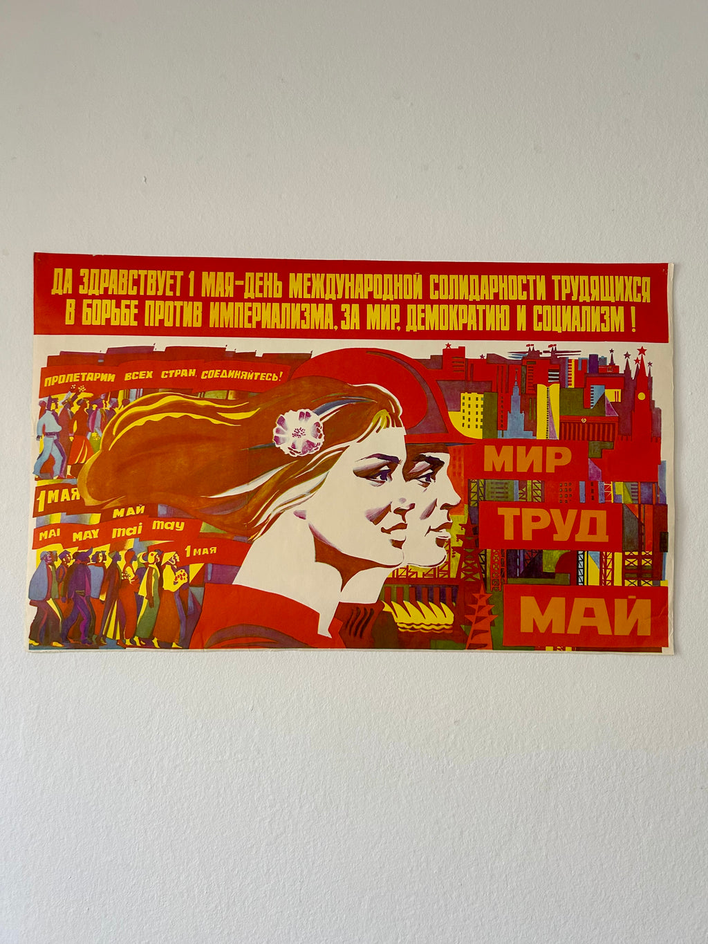 A vintage Soviet-style propaganda poster mounted on a white wall. The poster features a stylized illustration of a woman with flowing hair, bold red and yellow text, and a collage of industrial buildings and workers.