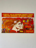 A vintage Soviet-style propaganda poster mounted on a white wall. The poster features a stylized illustration of a woman with flowing hair, bold red and yellow text, and a collage of industrial buildings and workers.