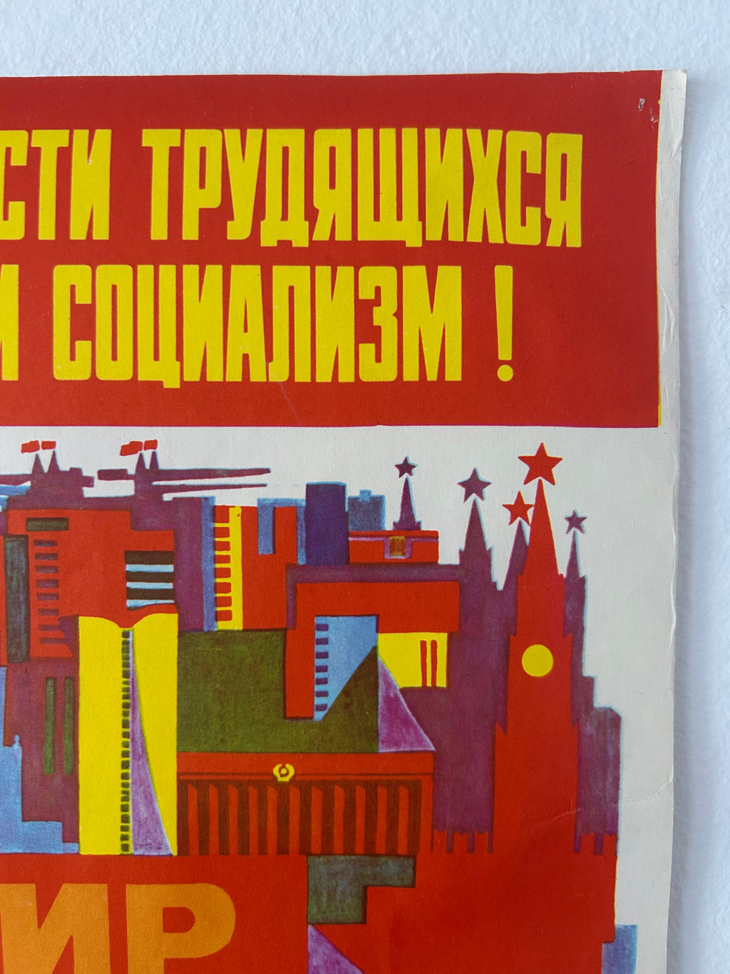 A close-up of a vintage Soviet propaganda poster with bold yellow Cyrillic text on a red background and a stylized multicolor cityscape illustration below, photographed against a light wall.