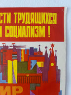 A close-up of a vintage Soviet propaganda poster with bold yellow Cyrillic text on a red background and a stylized multicolor cityscape illustration below, photographed against a light wall.