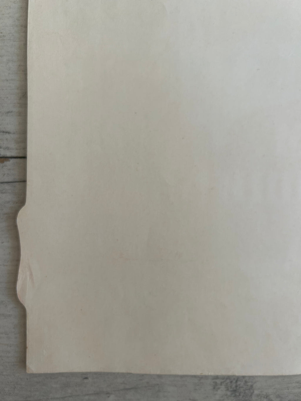 A blank, off-white sheet of paper or cardstock with a small folded tab on the left edge, resting on a gray wooden surface.
