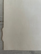 A blank, off-white sheet of paper or cardstock with a small folded tab on the left edge, resting on a gray wooden surface.