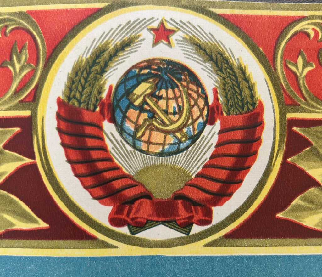 A close-up of a Soviet-style emblem printed on a textile or printed surface, featuring a red and gold wreath with wheat ears, a hammer and sickle over a globe, and a red star above, with a blue band at the bottom edge.