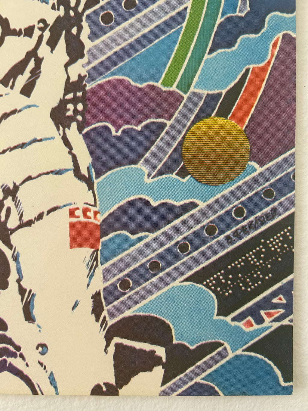 A close-up of a colorful graphic print or poster featuring abstract geometric shapes, stylized figures, and a gold circular element on a cream background.