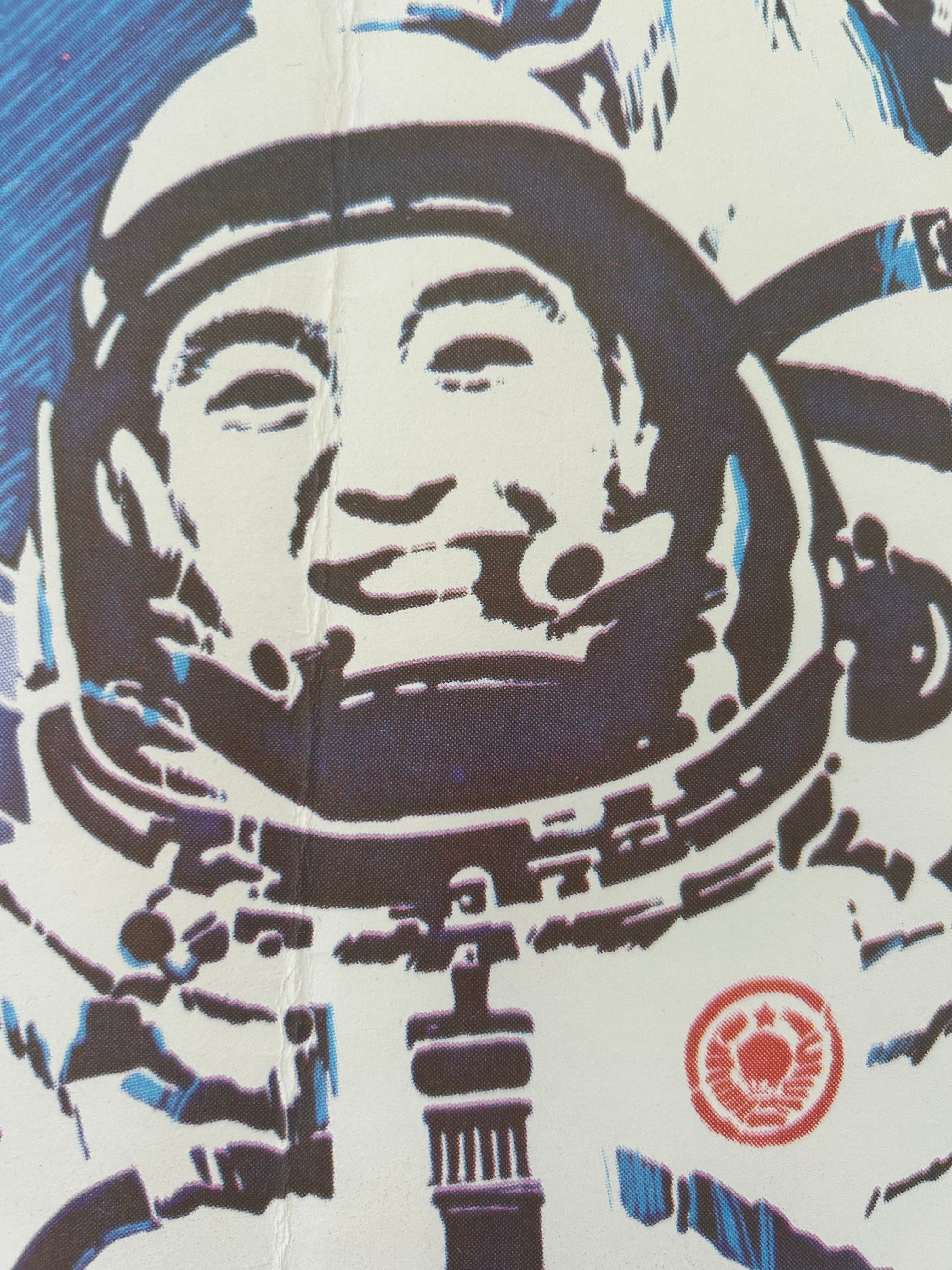 A close-up of a printed graphic showing part of an astronaut in a spacesuit, rendered in dark navy, white, and light blue with a small red circular emblem near the lower right.