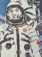 A close-up of a printed graphic featuring a stylized astronaut in a white spacesuit with bold black and blue linework, plus small red circular and square insignia on the chest and shoulder area.