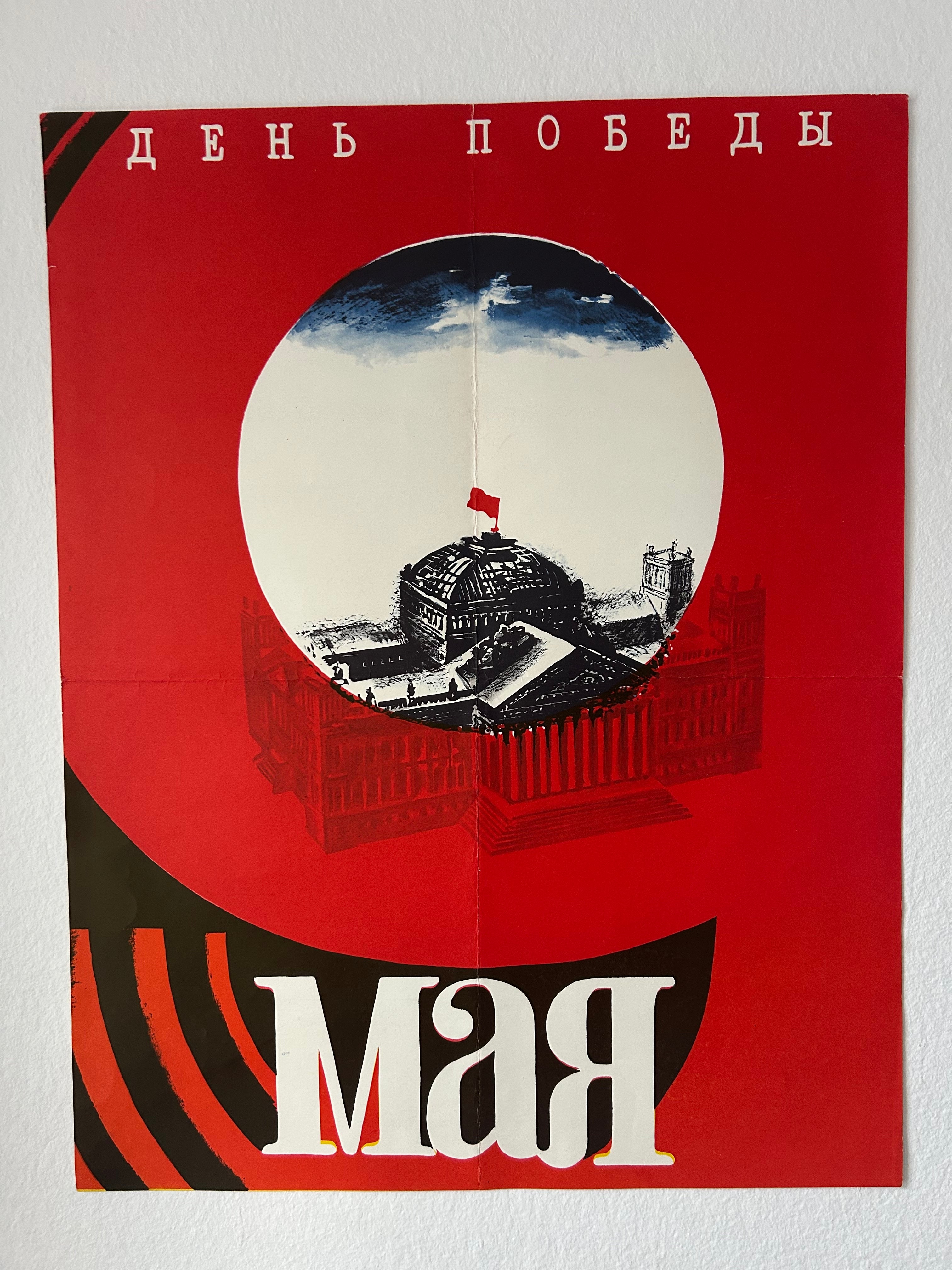 A vintage Soviet propaganda poster celebrating May Day, featuring a bold red background, a central circular illustration of a building with a flag, and the Cyrillic text 'ДЕНИК ПЕРИНИЙ' at the top and 'АМО' at the bottom.