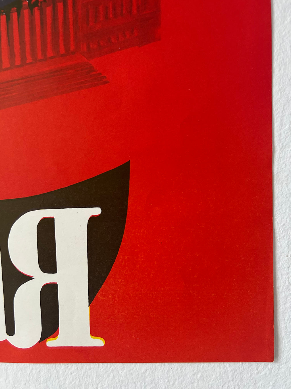 A close-up of a red poster or print featuring a large white serif letter 'R' over a dark brown shape, with part of a building illustration at the top and a white textured wall behind it.
