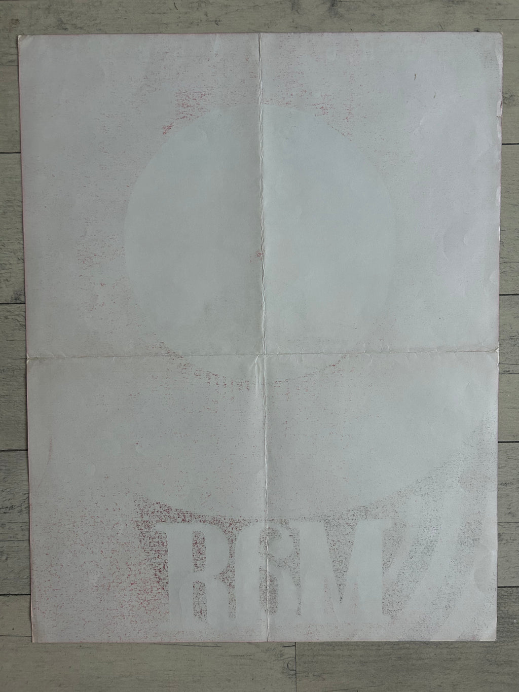 A square white sheet of paper with faint red speckled text and a faint circular graphic, laid flat on a gray wood plank surface.