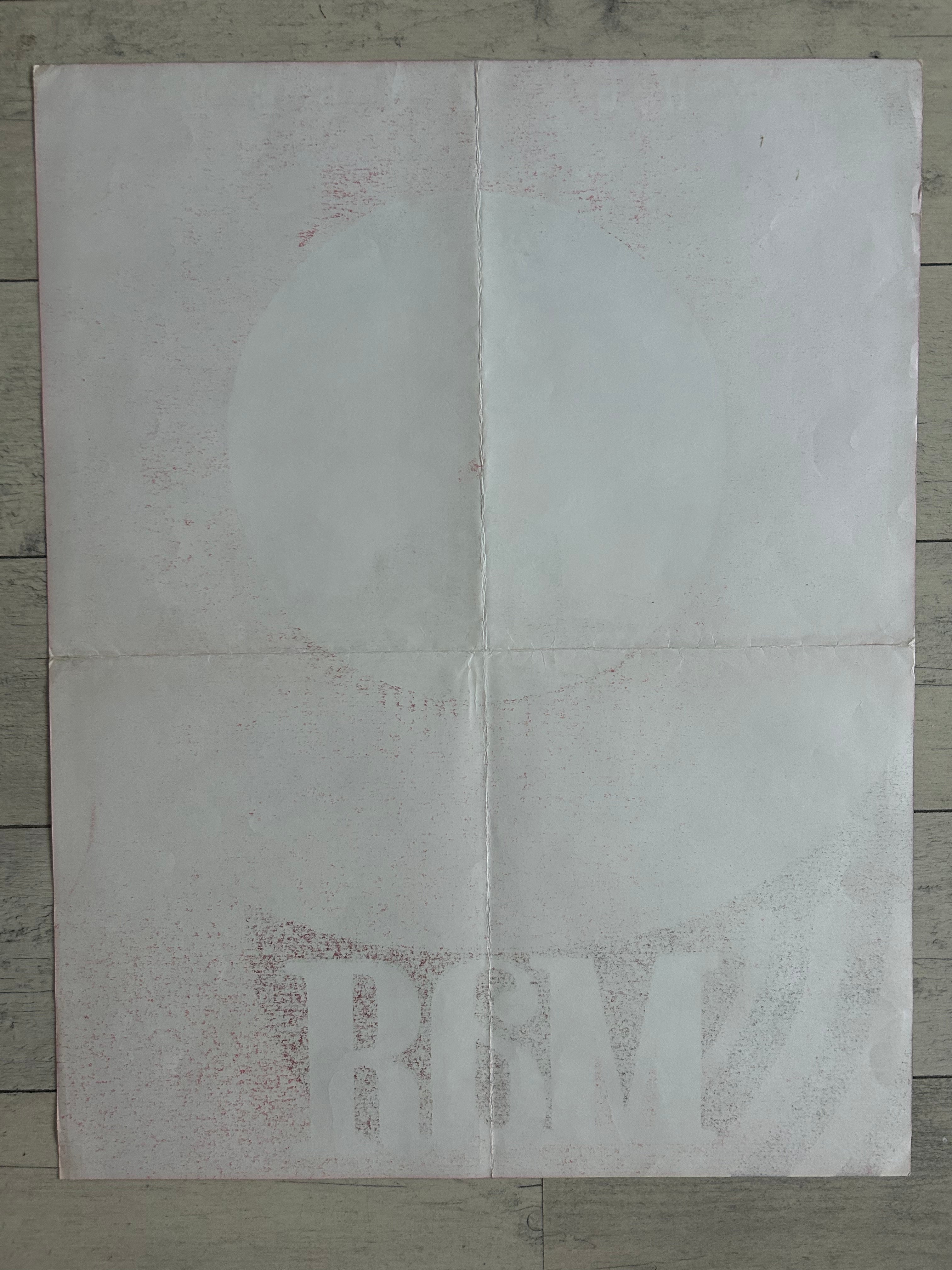 A square white sheet of paper with faint red speckled text and a faint circular graphic, laid flat on a gray wood plank surface.