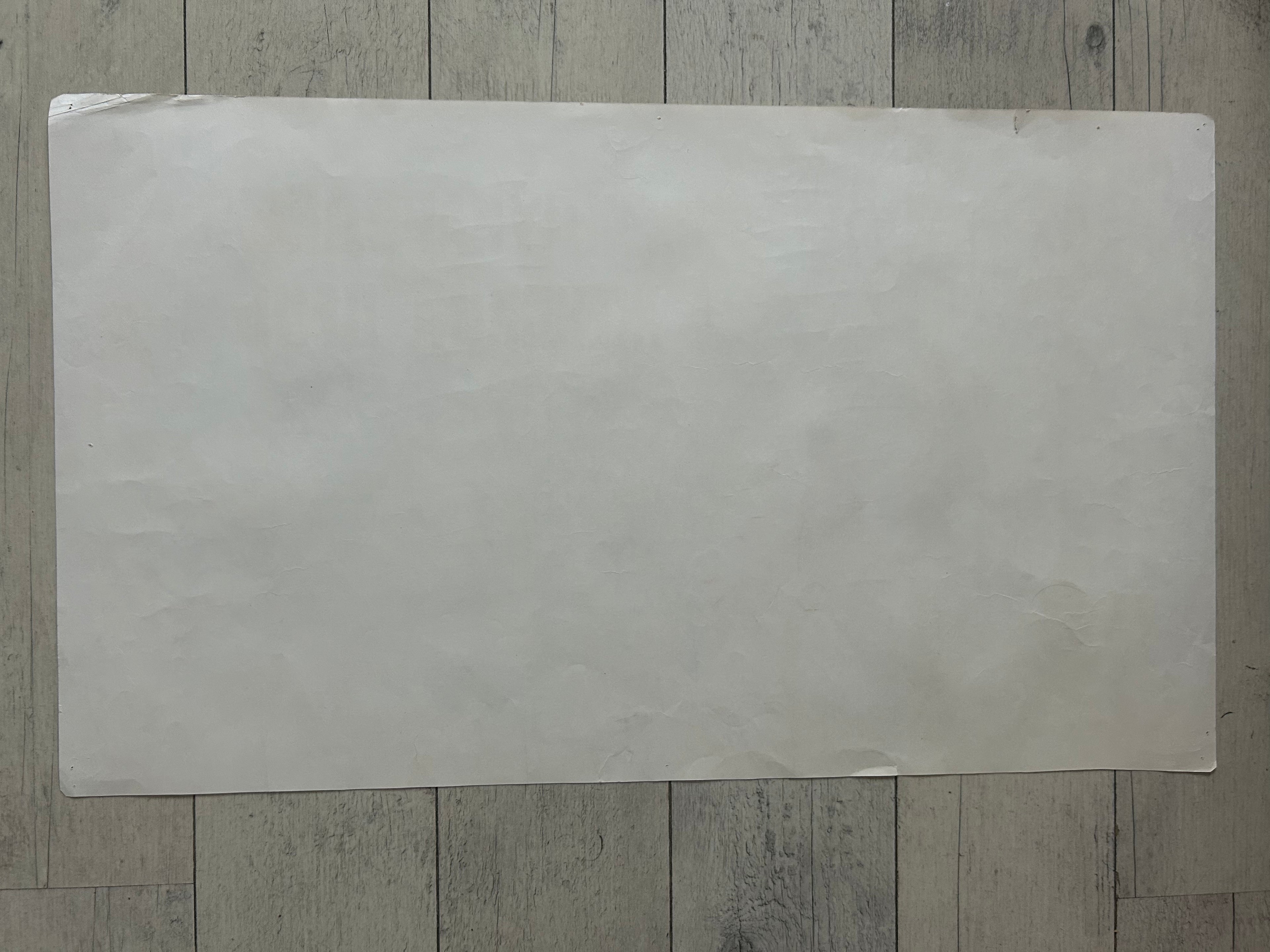 A large, blank, off-white rectangular sheet of paper or cardstock placed flat on a gray wood-plank floor.