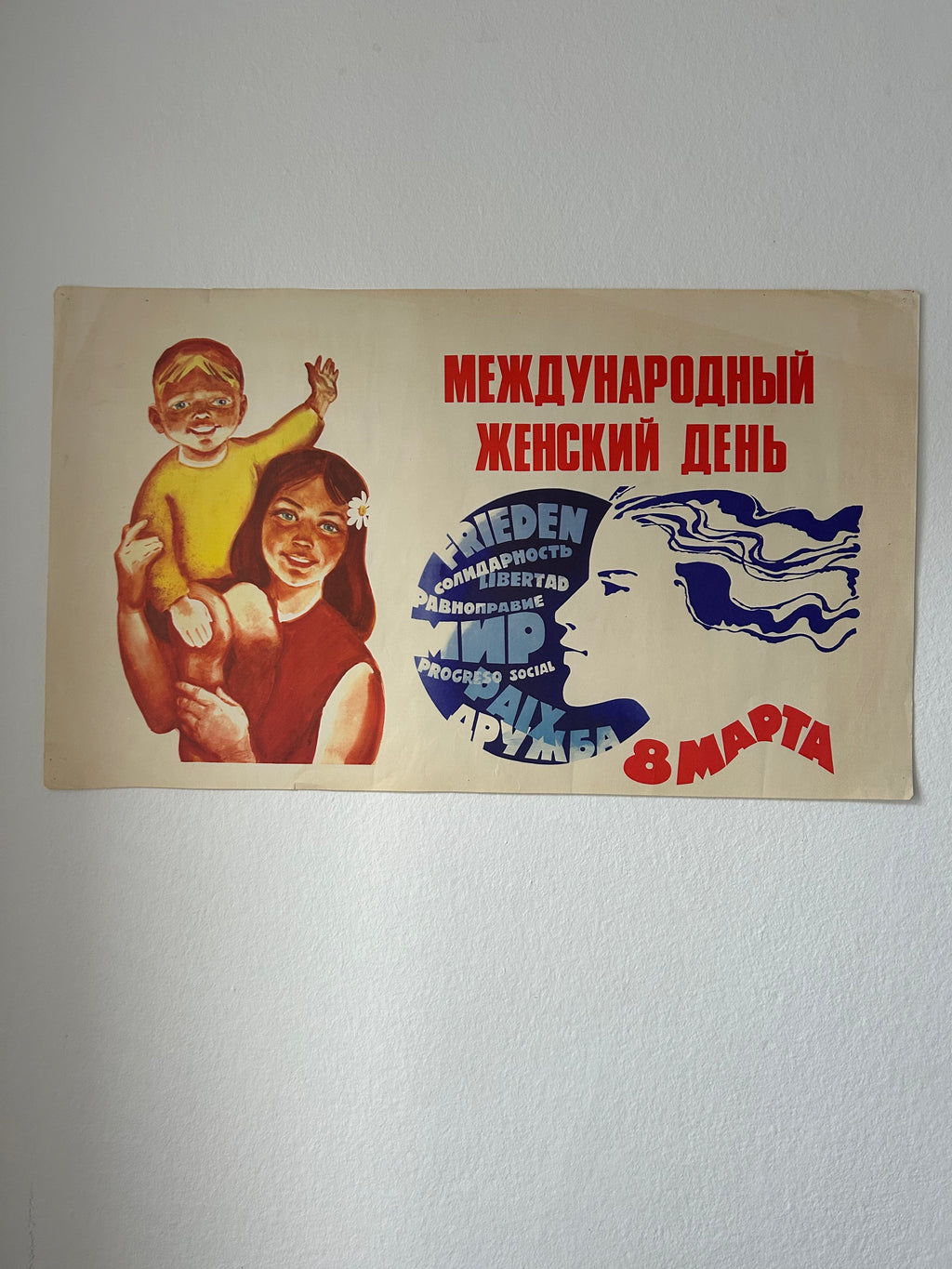 A vintage Soviet-era propaganda poster for International Women's Day (8 March) mounted on a white wall. The poster features a mother holding a child on the left, bold red Cyrillic text, and a blue stylized female profile with additional text on the right.