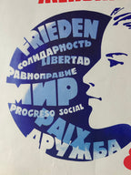A close-up of a printed graphic with a white face profile layered over large blue text blocks, including the words Frieden, Solidaridad, Libertad, Ravnopравие, MP, Progreso Social, and PaŪадание. The print appears on a light off-white background.