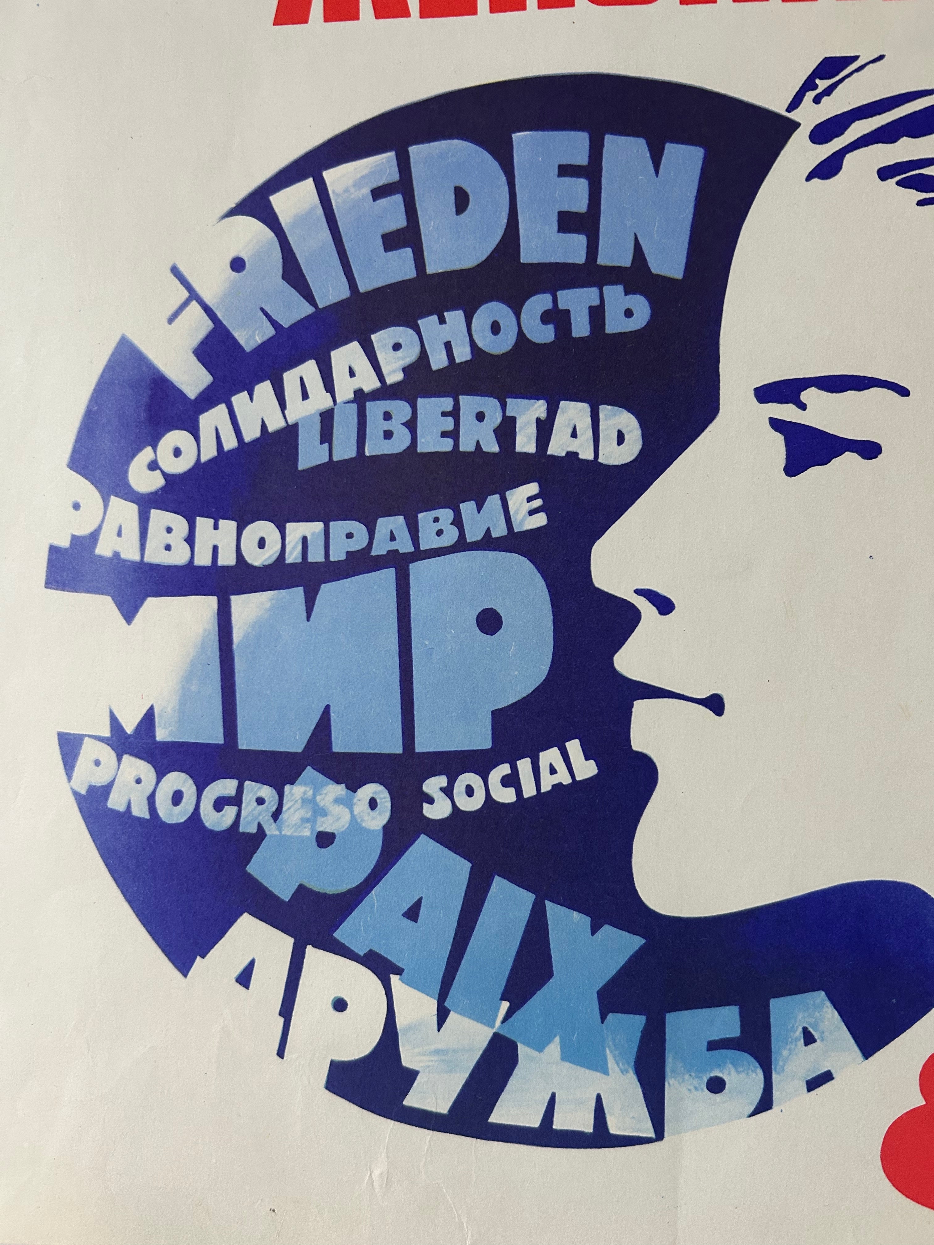 A close-up of a printed graphic with a white face profile layered over large blue text blocks, including the words Frieden, Solidaridad, Libertad, Ravnopравие, MP, Progreso Social, and PaŪадание. The print appears on a light off-white background.