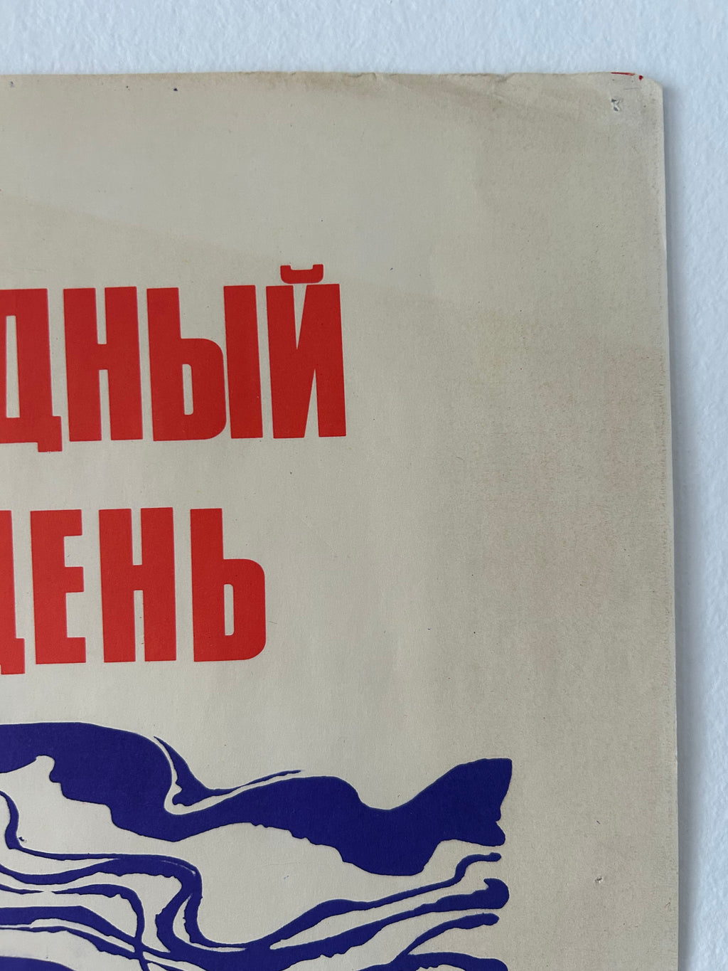 A close-up of a vintage Soviet-style poster or print mounted on a white wall. The beige paper shows bold red Cyrillic text at the top and a blue abstract wave-like graphic at the bottom.