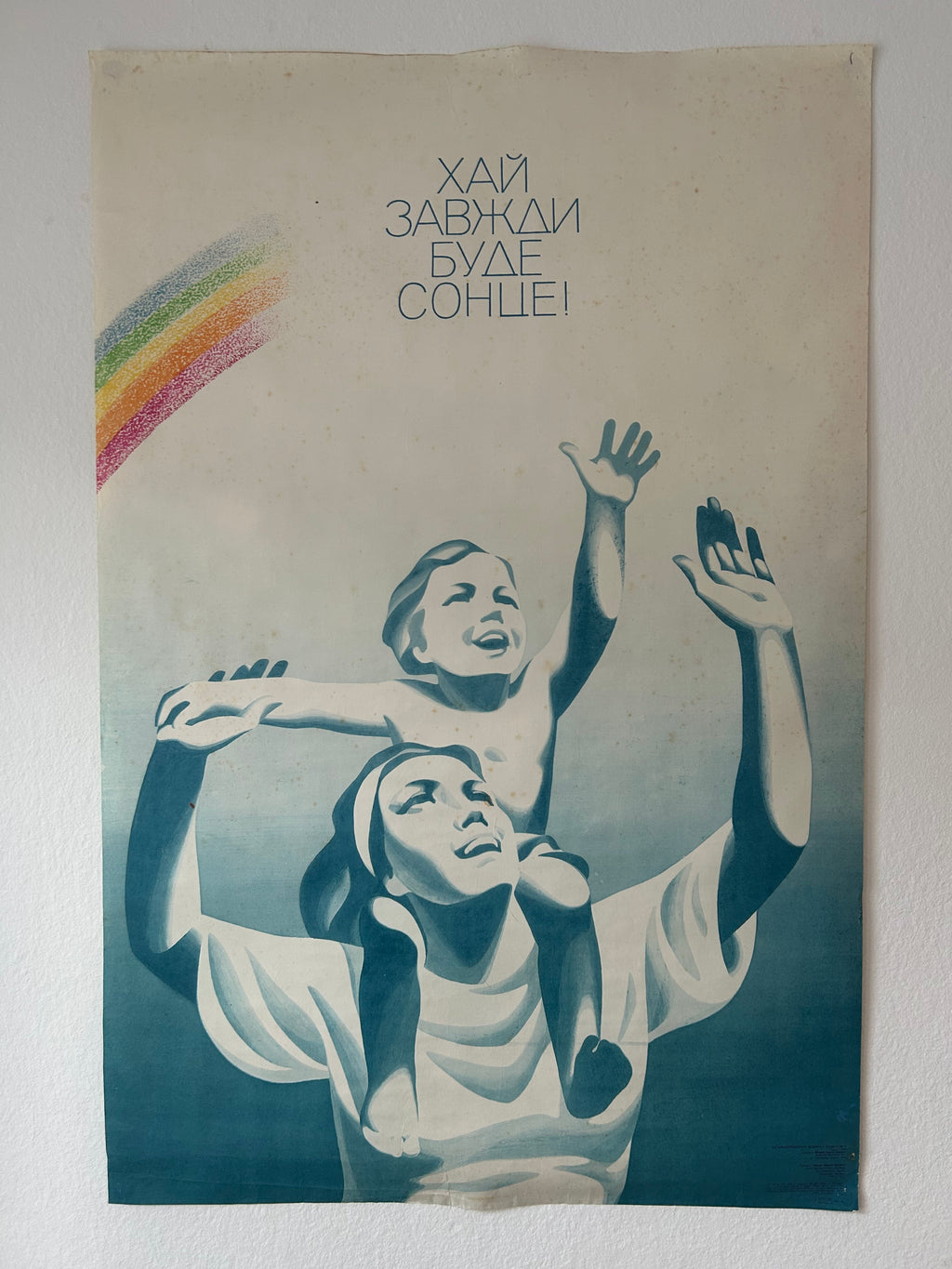 A vintage Soviet propaganda poster mounted on a white wall, featuring a mother and child with raised hands, a rainbow in the upper left, and the Russian text 'Кал здорим нежение здорима' at the top.