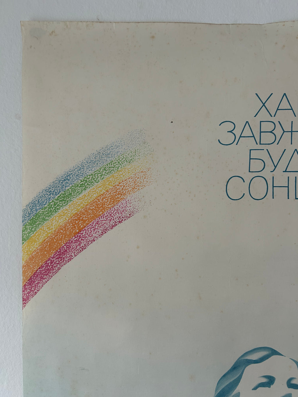 A close-up of a vintage-style printed card or poster with a cream background, a rainbow made of dotted colored stripes on the left, blue Cyrillic text on the right, and part of a blue illustrated face at the bottom.