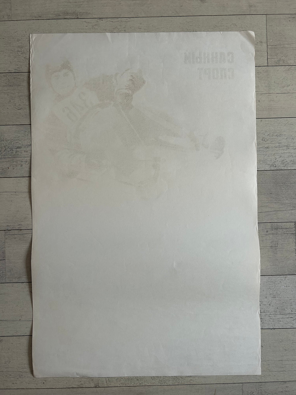 A large, blank off-white sheet of paper or poster lies flat on a gray wood-plank floor. Faint, barely legible text and a very low-contrast illustration are visible near the top right corner of the sheet.