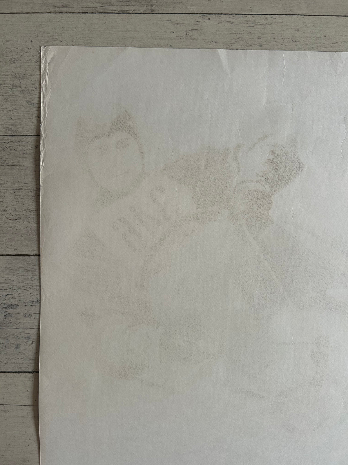 A square off-white art print or poster featuring a faint, low-contrast portrait sketch of a person in a baseball uniform, laid flat on a gray wood plank surface.