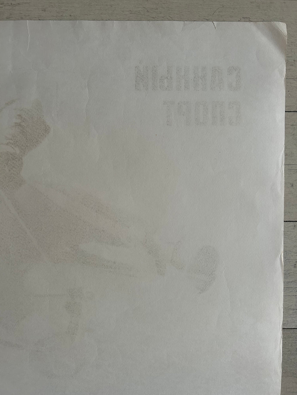 A square off-white paper sheet with faint, low-contrast printed text and a subtle illustration, photographed on a light wood surface.