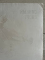 A square off-white paper sheet with faint, low-contrast printed text and a subtle illustration, photographed on a light wood surface.