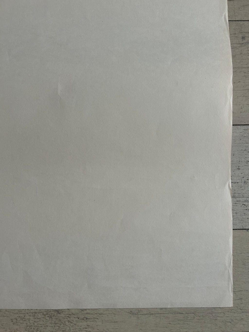 A large sheet of white or off-white paper laid flat on a light gray wood surface, filling most of the frame.