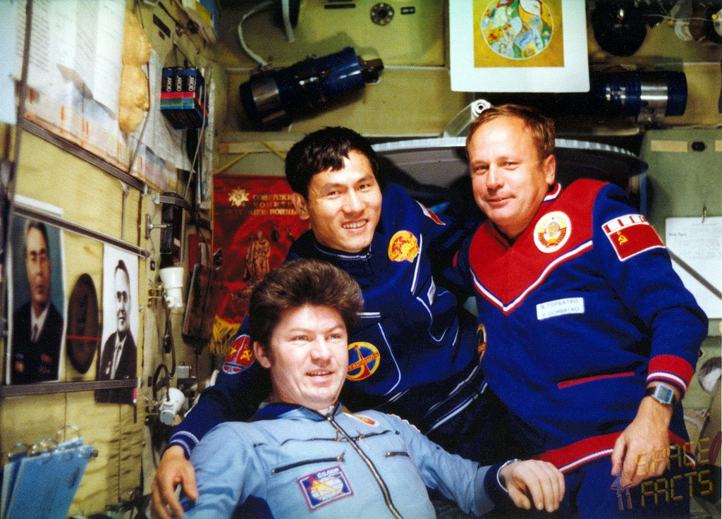 Three men in space-themed uniforms posing inside a cramped spacecraft or control room. The uniforms are blue and red with Soviet-era insignia, and the background is filled with equipment, posters, and wires.