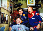 Three men in space-themed uniforms posing inside a cramped spacecraft or control room. The uniforms are blue and red with Soviet-era insignia, and the background is filled with equipment, posters, and wires.