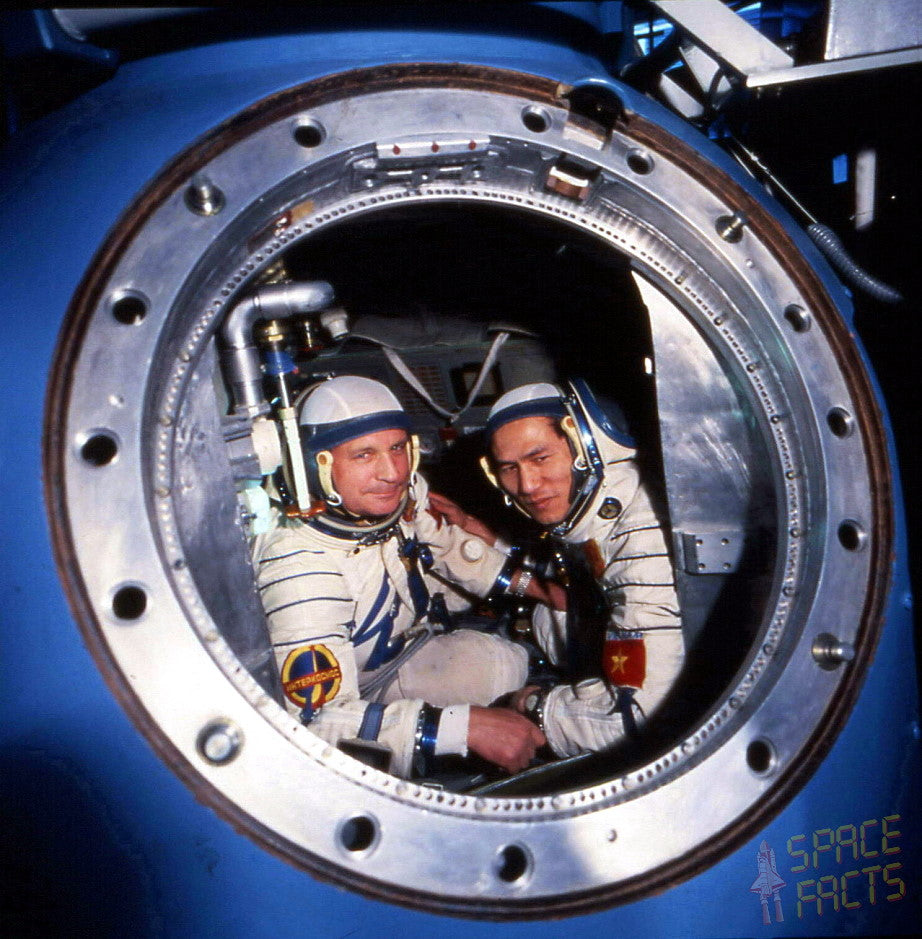 Two astronauts in white space suits look out through a circular window from inside a blue spacecraft or capsule. The photo is framed by the metallic rim of the window, with a small 'SPACE FACTS' mark in the lower right corner.