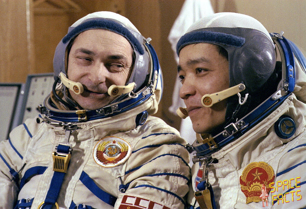 Two astronauts in white space suits with blue trim and helmets, seated indoors. One astronaut has a Soviet-era roundel patch on the chest, and the other has a red and gold emblem with the words 'SPACE FACTS'.