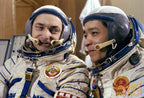 Two astronauts in white space suits with blue trim and helmets, seated indoors. One astronaut has a Soviet-era roundel patch on the chest, and the other has a red and gold emblem with the words 'SPACE FACTS'.